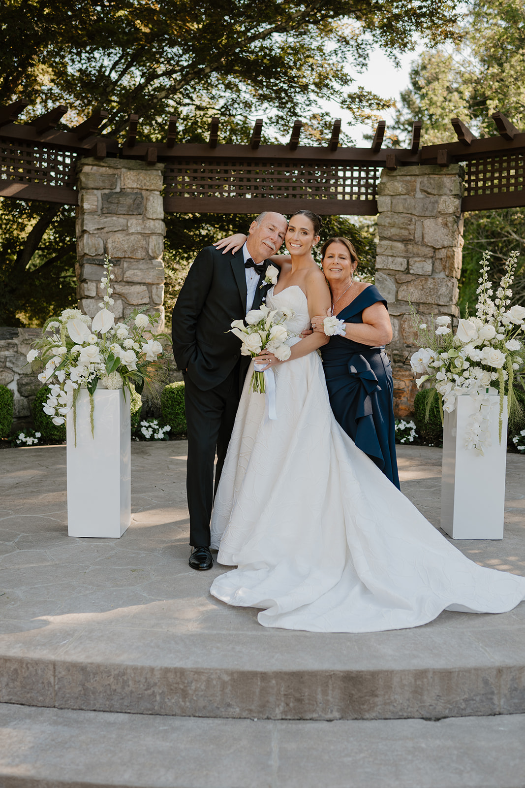 Bride posing with her smiling parents under an outdoor arbor framed by white floral pillars.